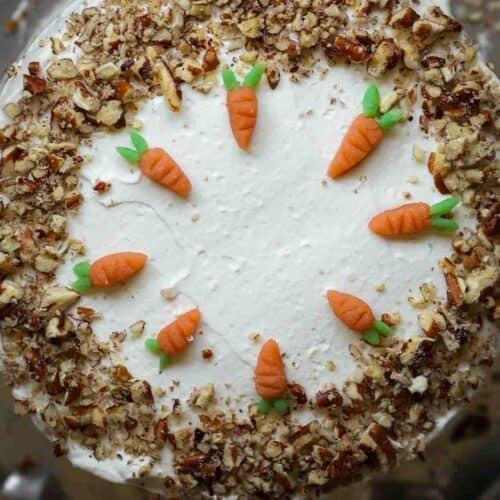 A round vegan carrot cake topped with white frosting, adorned with small fondant carrots in a circle on top. Chopped nuts line the edges, adding texture and contrast to the smooth icing.